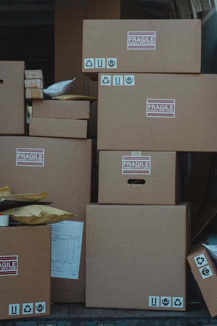 A collection of cardboard boxes of various sizes stacked on an outdoor pavement area, prepared for a home relocation. Some boxes are sealed with packing tape, and others have printed labels with handl
