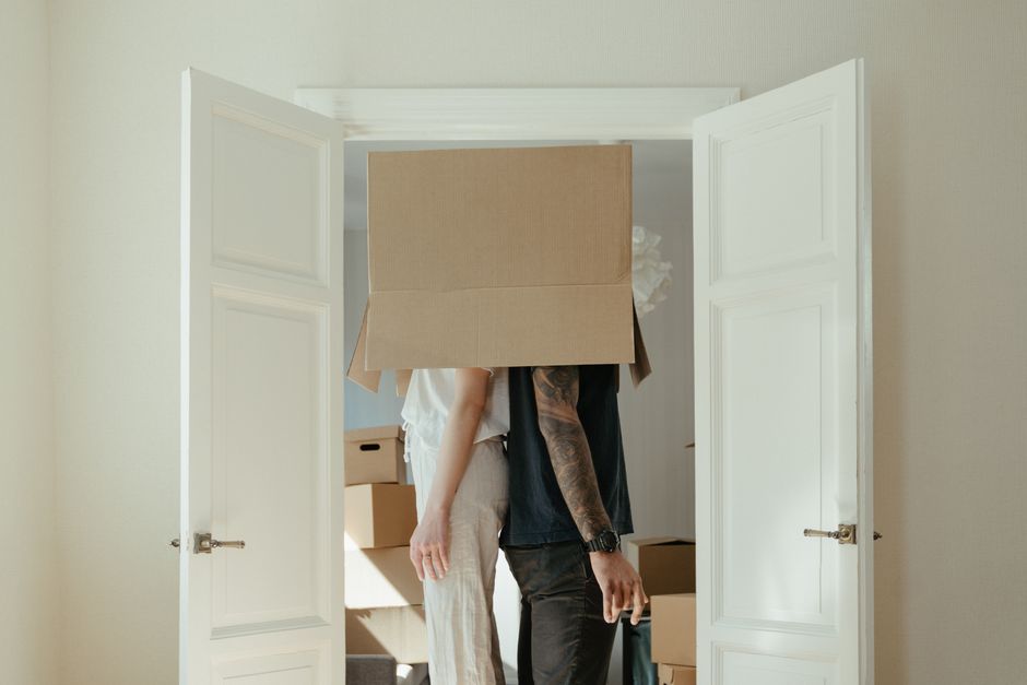 A person wearing dark trousers and grey socks standing on a wooden floor inside a home surrounded by several cardboard boxes used for packing during a house move. One box in the foreground has the wor