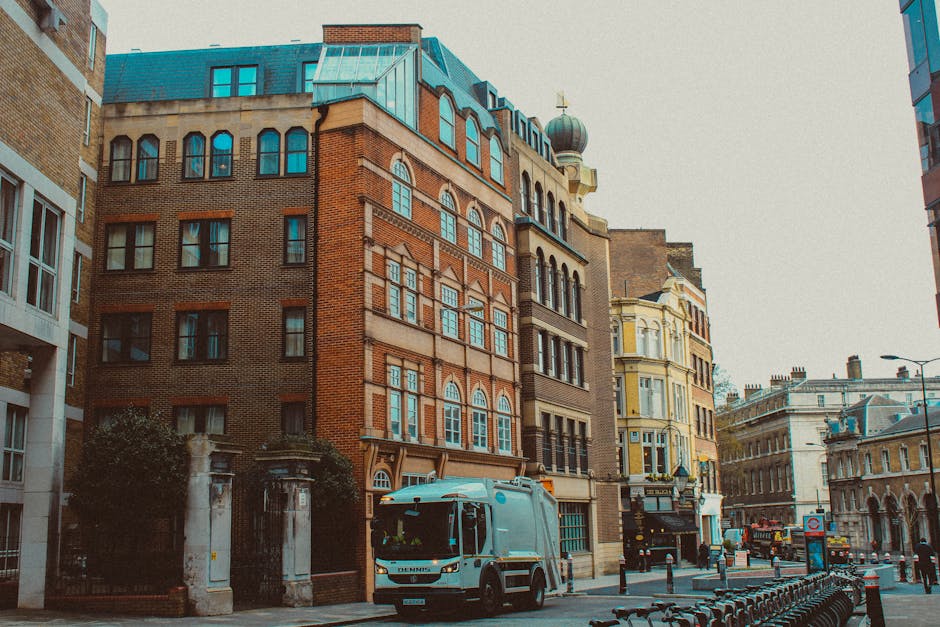 A street scene on Old Brompton Road featuring a white delivery van parked near a group of multi-storey buildings with varied architectural styles, including brick and ornate facades. The van, part of Man and Van Brompton's removals service, is positioned on the pavement adjacent to the entrance of a building, which has a stone gatepost and a wrought-iron fence. The van’s side door appears open, suggesting the loading or unloading process involved in home relocation or furniture transport. Nearby, large cardboard boxes and wrapped furniture items can be seen on the sidewalk, indicating packing and moving preparations. The surrounding area includes modern and historic buildings, with street lamps and bicycle racks visible along the road, under overcast sky lighting. This scene exemplifies urban residential removals, where careful handling of furniture, boxes, and packaging materials within a busy city environment is essential, supported by local removals specialists such as Man and Van Brompton.