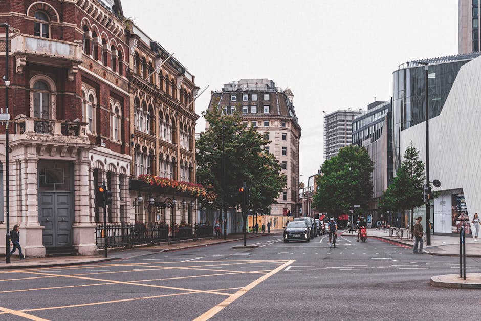 A street scene on Old Brompton Road featuring a white delivery van parked near a group of multi-storey buildings with varied architectural styles, including brick and ornate facades. The van, part of Man and Van Brompton's removals service, is positioned on the pavement adjacent to the entrance of a building, which has a stone gatepost and a wrought-iron fence. The van’s side door appears open, suggesting the loading or unloading process involved in home relocation or furniture transport. Nearby, large cardboard boxes and wrapped furniture items can be seen on the sidewalk, indicating packing and moving preparations. The surrounding area includes modern and historic buildings, with street lamps and bicycle racks visible along the road, under overcast sky lighting. This scene exemplifies urban residential removals, where careful handling of furniture, boxes, and packaging materials within a busy city environment is essential, supported by local removals specialists such as Man and Van Brompton.