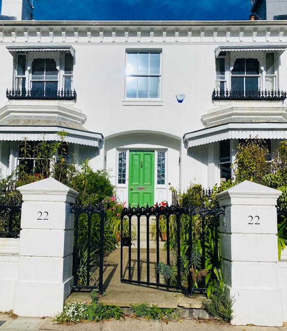 The image depicts the front façade of a white, two-story residential building with a central bright green front door surrounded by glass panes. Flanking the front door are two symmetrical bay windows with black wrought iron balcony railings on the upper level. The house has decorative white cornices along the roofline and a large, evenly spaced rectangular window on the upper floor. In front of the property, a black metal gate is partially open, connected to white stone pillars numbered '22', which form a small front garden area. Inside the garden, various green plants, shrubs, and flowering plants are visible, with some arranged along the pathway leading to the front door. The scene suggests a typical home in Brompton, with the context of house removals or moving services, as the image is associated with Man and Van Brompton and their same-day relocation offerings. The environment appears well-lit, indicating daytime, and the overall setting emphasizes the interior and exterior elements involved in move-related logistics.